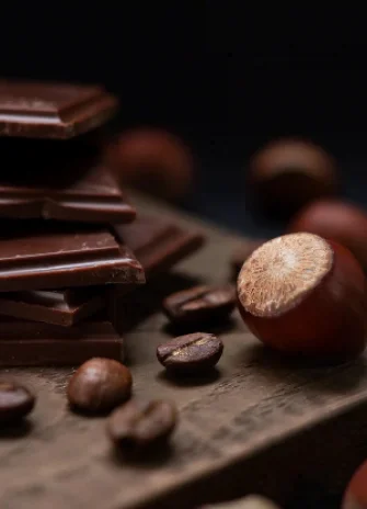 Pieces of dark chocolate on a wooden surface next to hazelnuts and coffee beans.